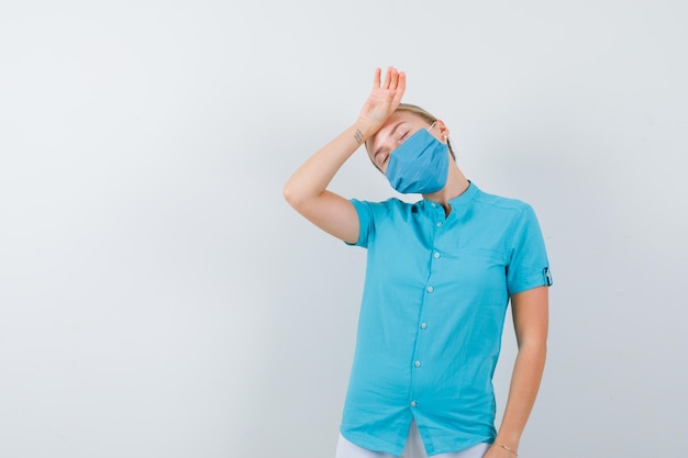 Junge Ärztin, die Hand auf der Stirn in medizinischer Uniform hält, Maske und müde aussieht