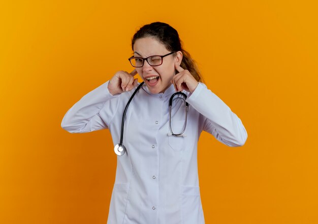 Junge Ärztin, die medizinische Robe und Stethoskop mit den geschlossenen Ohren der Brille lokalisiert trägt