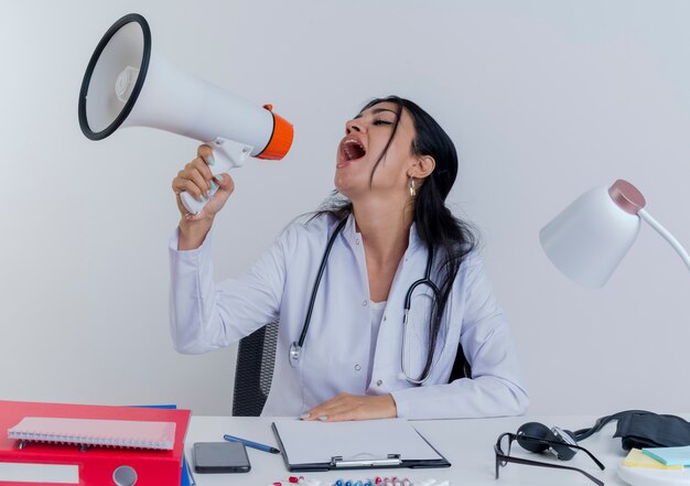 Junge Ärztin, die medizinische Robe und Stethoskop trägt, sitzt am Schreibtisch mit medizinischen Werkzeugen, die Hand auf Schreibtisch setzen, die durch Sprecher lokalisiert betrachten und sprechen