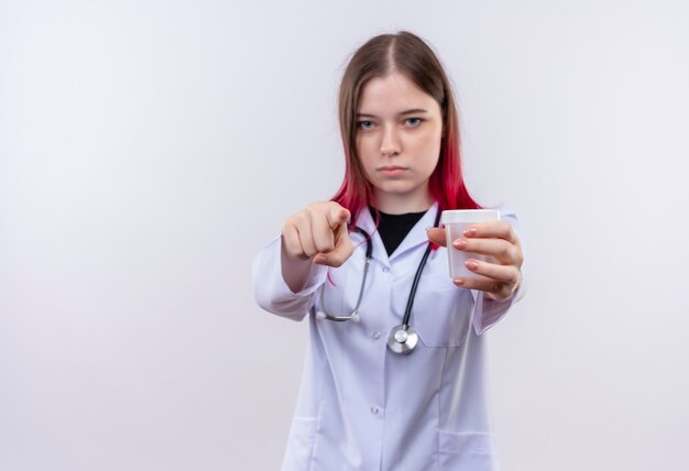 junge Ärztin, die medizinisches Gewand des Stethoskops trägt, das leere Dose heraushält, die Sie Geste auf isolierter weißer Wand mit Kopienraum zeigt