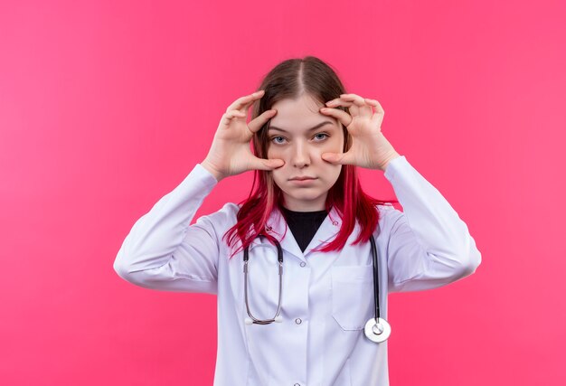 junge Ärztin, die medizinisches Kleid des Stethoskops trägt, hält ihre Augen offen mit Händen auf rosa isolierter Wand