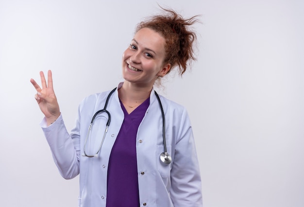 Junge Ärztin, die weißen Kittel mit Stethoskop trägt, zeigt und zeigt mit den Fingern Nummer drei, die fröhlich über weißer Wand stehend lächeln