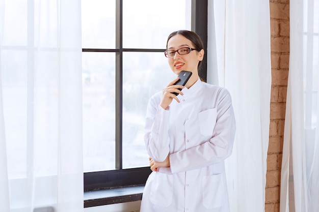 Junge Ärztin im Laborkittel, die Handy hält, während sie in der Nähe des Fensters steht.