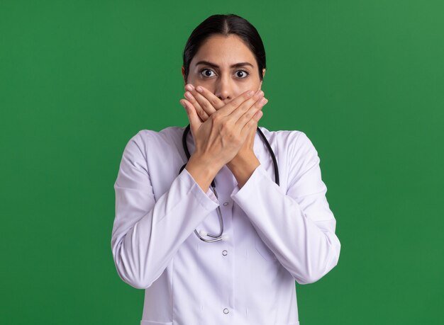 Junge Ärztin im medizinischen Mantel mit dem Stethoskop, das Front betrachtet, das schockiert wird, den Mund mit den Händen stehend über grüner Wand bedeckt