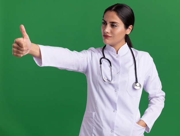 Junge Ärztin im medizinischen Mantel mit Stethoskop, das mit ernstem selbstbewusstem Ausdruck beiseite schaut und Daumen oben zeigt über grüner Wand