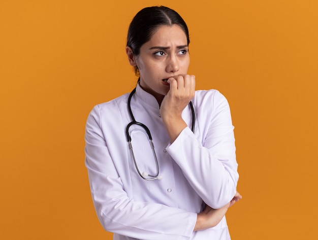 Junge Ärztin im medizinischen Mantel mit Stethoskop um ihren Hals, der gestresste und nervöse beißende Nägel beiseite schaut, die über orange Wand stehen