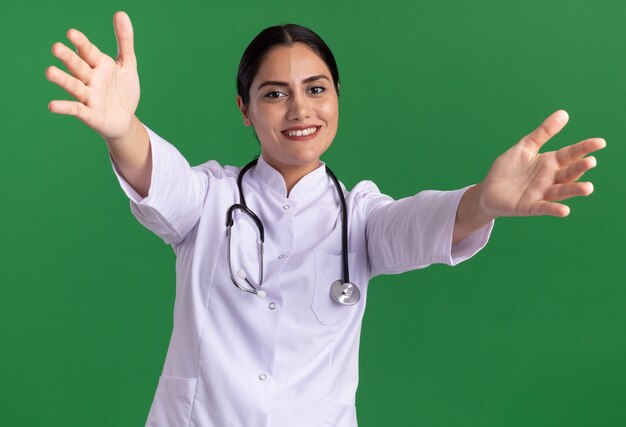 Junge Ärztin im medizinischen Mantel mit Stethoskop um ihren Hals, der vorne lächelnd mit glücklichem Gesicht schaut, das Begrüßungsgeste mit weit öffnenden Händen der Hände macht, die über grüner Wand stehen