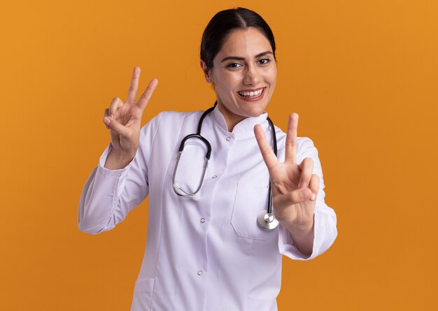 Junge Ärztin im medizinischen Mantel mit Stethoskop um ihren Hals, der vorne mit dem glücklichen Gesicht lächelnd zeigt, das V-Zeichen zeigt, das über orange Wand steht