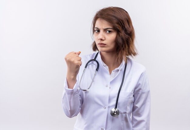 Junge Ärztin im weißen Kittel mit der geballten Faust des Stethoskops, die Kamera mit wütendem Gesicht betrachtet