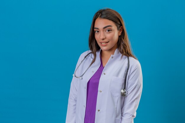 Junge Ärztin im weißen Kittel mit lächelndem Phonendoskop, das Kamera über lokalisiertem blauem Hintergrund betrachtet