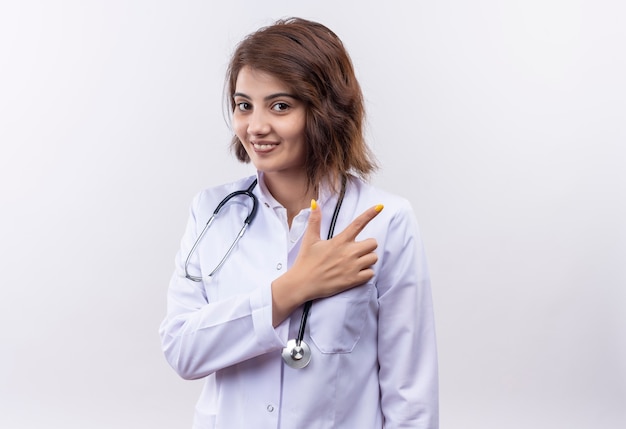 Junge Ärztin im weißen Kittel mit lächelndem Stethoskop, das fröhlich mit Zeigefinger auf die Seite zeigt, die über weißer Wand steht