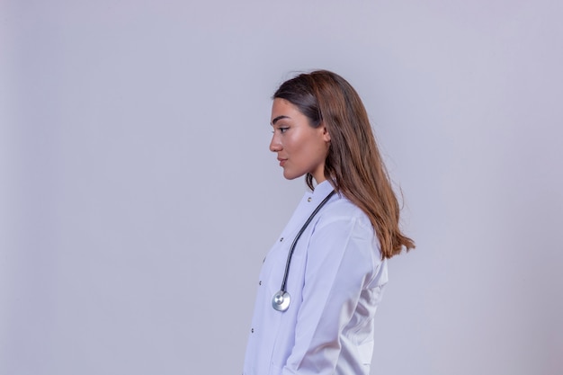Junge Ärztin im weißen Kittel mit Phonendoskop, das zur Seitenprofilhaltung steht, die über weißem lokalisiertem Hintergrund steht