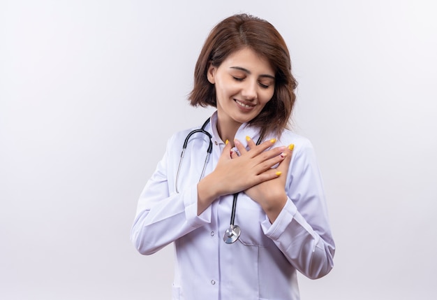 Junge Ärztin im weißen Kittel mit Stethoskop, das gekreuzte Arme über Brust hält, die positive Emotionen fühlen