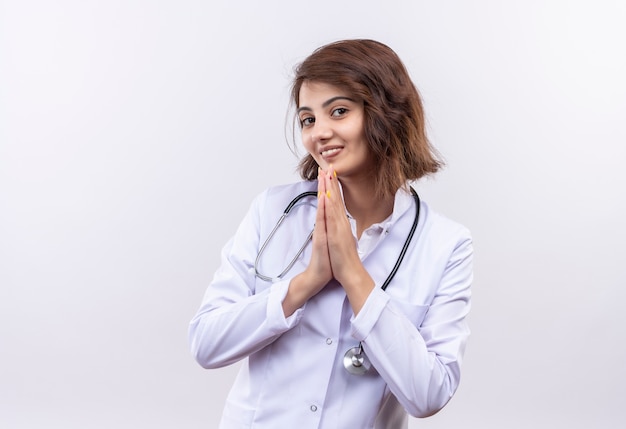 Junge Ärztin im weißen Kittel mit Stethoskop, das Hände in Gebetsgeste hält, dankbar und glücklich fühlt