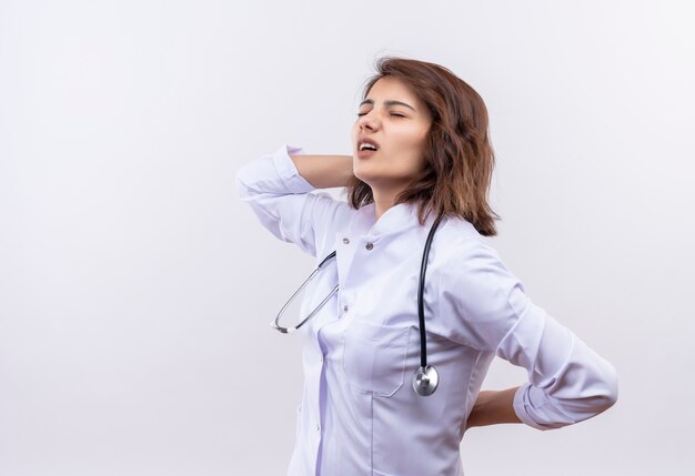 Junge Ärztin im weißen Kittel mit Stethoskop, das ihren Rücken berührt, der Schmerz fühlt, der über weißer Wand steht