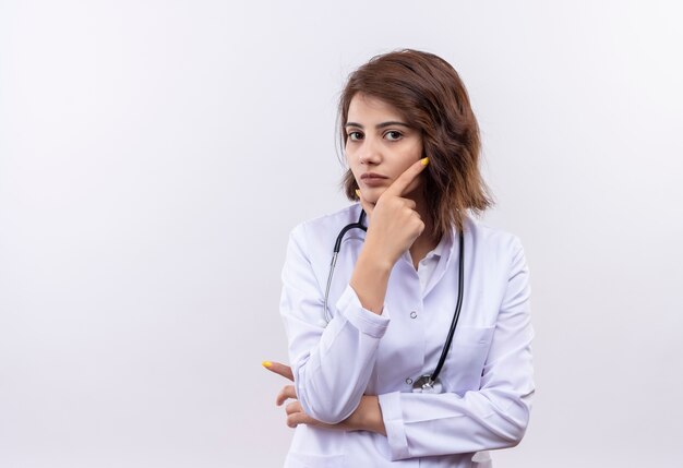 Junge Ärztin im weißen Kittel mit Stethoskop, das mit Hand auf Kinn mit nachdenklichem Ausdruck auf Gesichtsdenken beiseite schaut