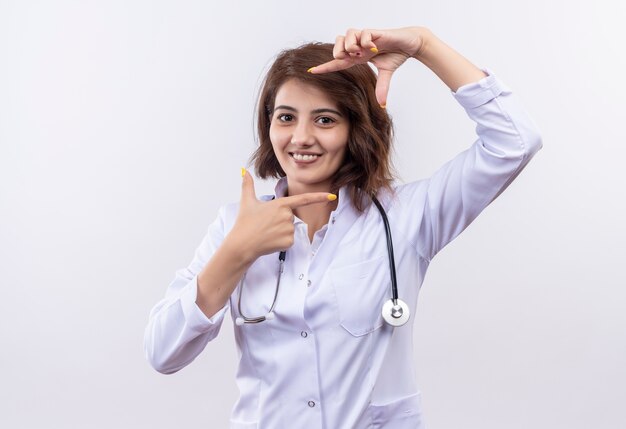 Junge Ärztin im weißen Kittel mit Stethoskop, das Rahmen mit den Fingern macht, die durch Finger schauen, die breit über weißer Wand stehend lächeln