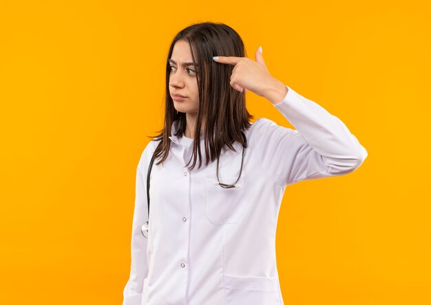 Junge Ärztin im weißen Kittel mit Stethoskop um den Hals, das mit dem Finger auf ihre Schläfe zeigt, konzentrierte sich auf eine Aufgabe, die über orange Wand steht