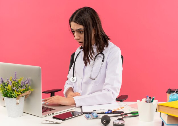 Junge Ärztin im weißen Kittel mit Stethoskop um ihren Hals, der am Laptop arbeitet, der verwirrt am Tisch über rosa Wand sitzt