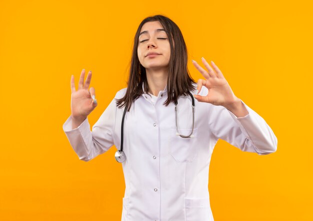 Junge Ärztin im weißen Kittel mit Stethoskop um ihren Hals, der sich mit geschlossenen Augen entspannt und Meditationsgeste mit Fingern macht, die über orange Wand stehen
