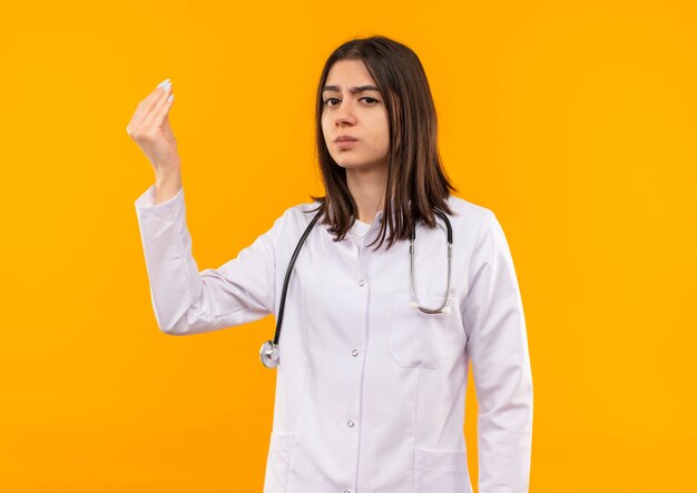 Junge Ärztin im weißen Kittel mit Stethoskop um ihren Hals, die nach vorne unzufrieden gestikulierend mit Hand steht, die über orange Wand steht
