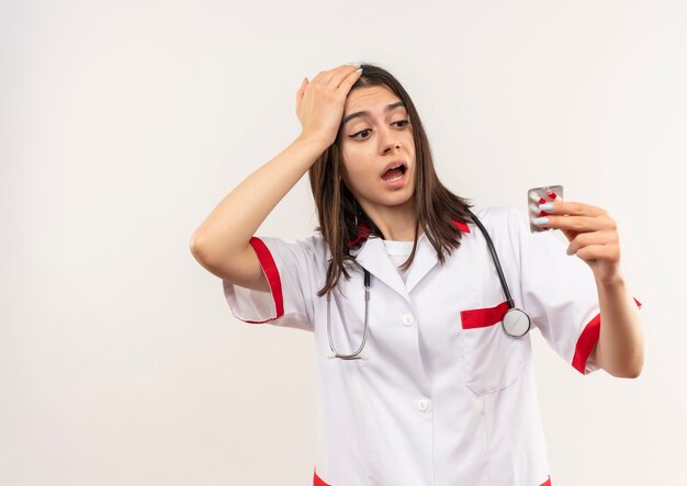 Junge Ärztin im weißen Kittel mit Stethoskop um ihren Hals hält Blister mit Pillen, die es betrachten, verwirrt über weißer Wand stehend