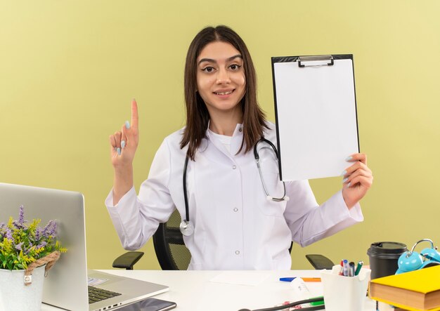 Junge Ärztin im weißen Kittel mit Stethoskop um ihren Hals hält Zwischenablage, die Zeigefinger zeigt, der zuversichtlich am Tisch mit Laptop über Lichtwand sitzt