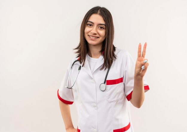 Junge Ärztin im weißen Kittel mit Stethoskop um ihren Hals zeigt und zeigt mit den Fingern Nummer zwei lächelnd über weißer Wand stehend
