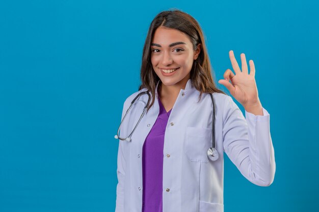 Junge Ärztin im weißen Mantel mit dem lächelnden Phonendoskop, das ok Zeichen mit den Fingern über lokalisiertem blauem Hintergrund tut