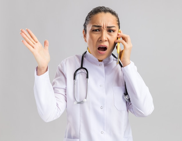 Junge Ärztin im weißen medizinischen Kittel mit Stethoskop um den Hals, die genervt und irritiert aussieht, während sie mit dem Handy spricht