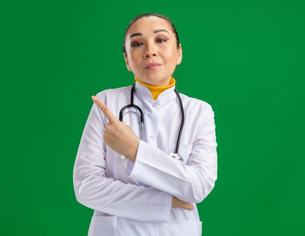 Junge Ärztin im weißen Medizinkittel mit Stethoskop um den Hals, die selbstbewusst aussieht und mit dem Zeigefinger auf die Seite zeigt, die über der grünen Wand steht