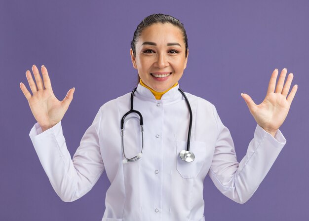Junge Ärztin im weißen Medizinmantel mit Stethoskop um den Hals, die die Arme hebt und fröhlich über lila Wand steht?