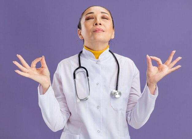 Junge Ärztin im weißen Medizinmantel mit Stethoskop um den Hals, die Meditationsgeste mit Fingern mit geschlossenen Augen macht, die sich über lila Wand entspannen?