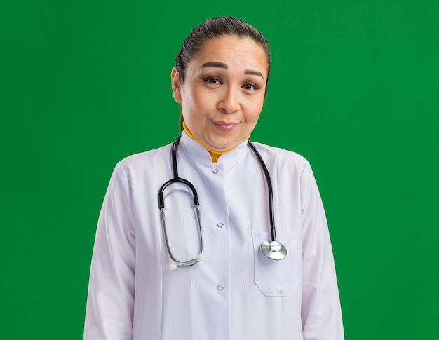 Junge Ärztin im weißen Medizinmantel mit Stethoskop um den Hals mit schüchternem Lächeln im Gesicht, das über grüner Wand steht
