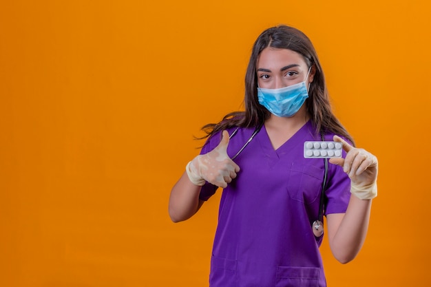 Junge Ärztin in der medizinischen Uniform mit Phonendoskop, das Schutzmaske und Handschuhe trägt, die Blister mit Pillen lächeln, die Daumen oben über lokalisiertem orangefarbenem Hintergrund zeigen