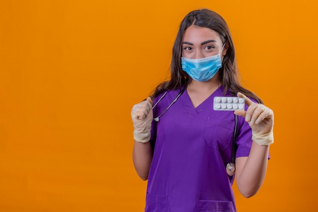 Junge Ärztin in der medizinischen Uniform mit Phonendoskop, das Schutzmaske und Handschuhe trägt, die Blister mit Pillen und erhabener Faust über lokalisiertem orangefarbenem Hintergrund lächeln