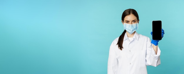 Junge Ärztin in medizinischer Gesichtsmaske und Krankenhausuniform mit Handy-App-Bildschirminterfa