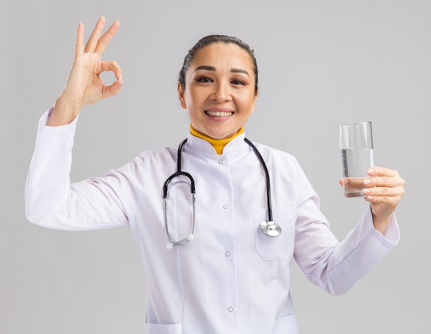 Junge Ärztin in weißem medizinischem Kittel mit Stethoskop um den Hals, die ein Glas Wasser hält und lächelt, das ein ok-Zeichen über der weißen Wand zeigt