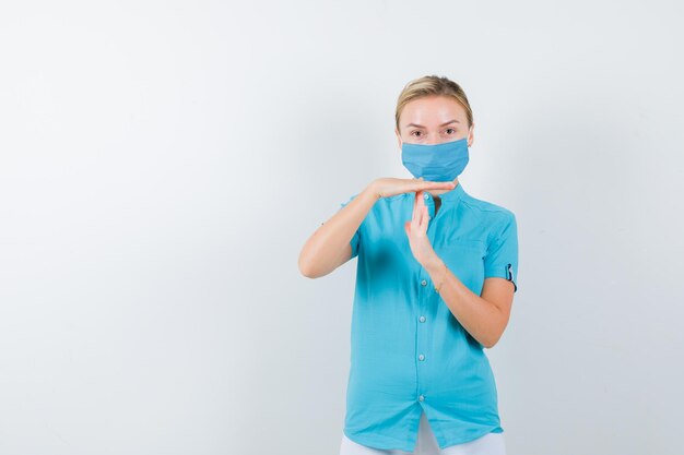 Junge Ärztin zeigt Timeout-Geste in medizinischer Uniform, Maske und sieht selbstbewusst aus