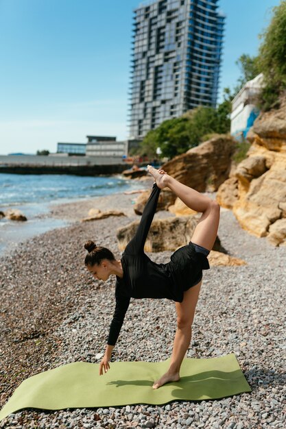 Junge schlanke Frau, die Übung für Muskel auf Yogamatte im Freien am Kieselstrand durch das Meer tut