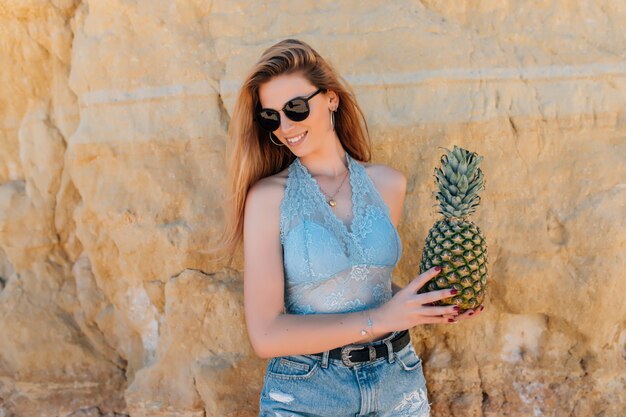 Junge schlanke Frau im Bikini und in der Sonnenbrille, die frische Ananas nahe dem Meer halten