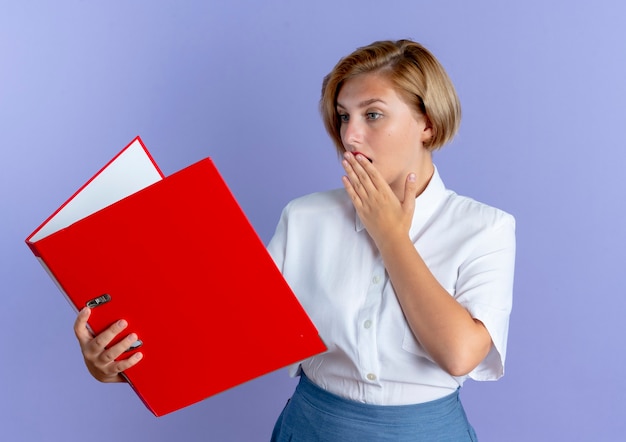 Junge schockierte blonde russische Mädchen hält und schaut auf Datei Ordner legt Hand auf Mund isoliert auf lila Hintergrund mit Kopie Raum