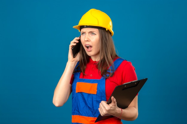 Junge schockierte Frau in der Bauuniform und im gelben Sicherheitshelm, der Zwischenablage hält und durch Smartphone steht auf blauem Hintergrund spricht