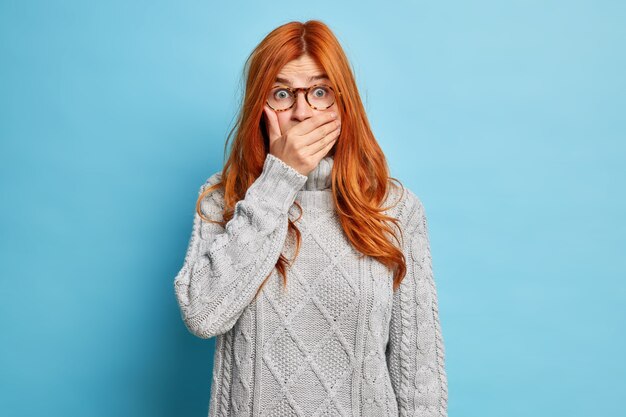 Junge schockierte rothaarige junge Frau bedeckt Mund und starrt verblüfft hört peinliche Nachrichten trägt transparente Brille grauer Strickpullover.