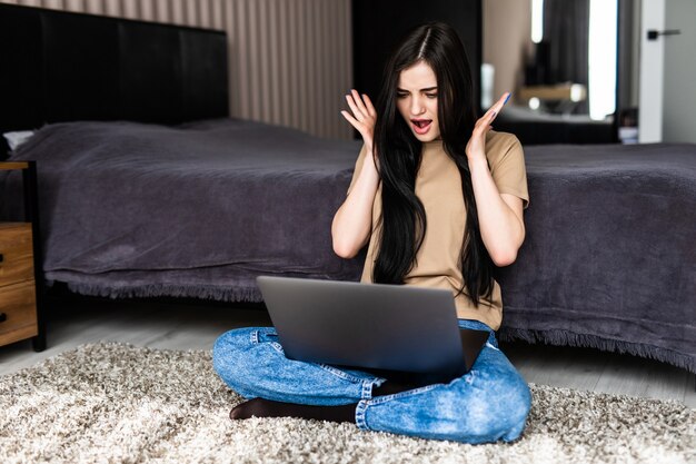 Junge schockierte überraschte junge Frau mit Videoanruf über Laptop-Computer auf dem Boden liegend im gemütlichen Wohnzimmer während der Fernarbeit von zu Hause aus
