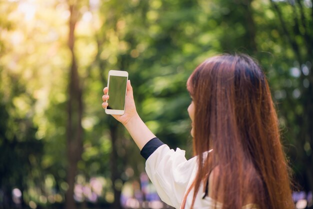 Junge schöne asiatische Frauen mit langen braunen Haaren ein Selfie auf ihrem Handy im Park. Natürliche Beleuchtung, lebendige Farben.