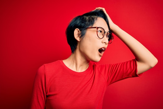 Junge schöne asiatische Mädchen trägt lässiges T-Shirt und Brille über isoliertem rotem Hintergrund überrascht mit der Hand auf dem Kopf wegen Fehler erinnern Fehler vergessen schlechtes Gedächtniskonzept vergessen