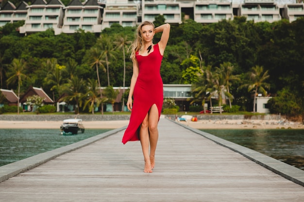 Junge schöne attraktive Frau, die allein auf Pier im Luxusresorthotel, Sommerferien, rotes langes Kleid, blondes Haar, sexy Kleidung, tropischer Strand, verführerisch, sinnlich, lächelnd steht