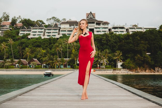 Junge schöne attraktive Frau, die allein auf Pier im Luxusresorthotel, Sommerferien, rotes langes Kleid, blondes Haar, sexy Kleidung, tropischer Strand, verführerisch, sinnlich, lächelnd steht