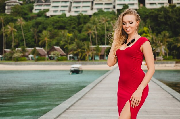 Junge schöne attraktive Frau, die allein auf Pier im Luxusresorthotel, Sommerferien, rotes langes Kleid, blondes Haar, sexy Kleidung, tropischer Strand, verführerisch, sinnlich, lächelnd steht